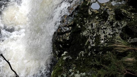 Slow pan up over waterfall pouring rapidly over brown moss covered rocks with puddles in natural, rugged wilderness setting creating splash and bubbles in river. Slow motion footage.