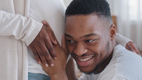 Close-up male head loving caring afro american black ethnic husband father hugs embraces touching listening pregnant female belly enjoying sounds of unborn future baby feeling happy, pregnancy concept