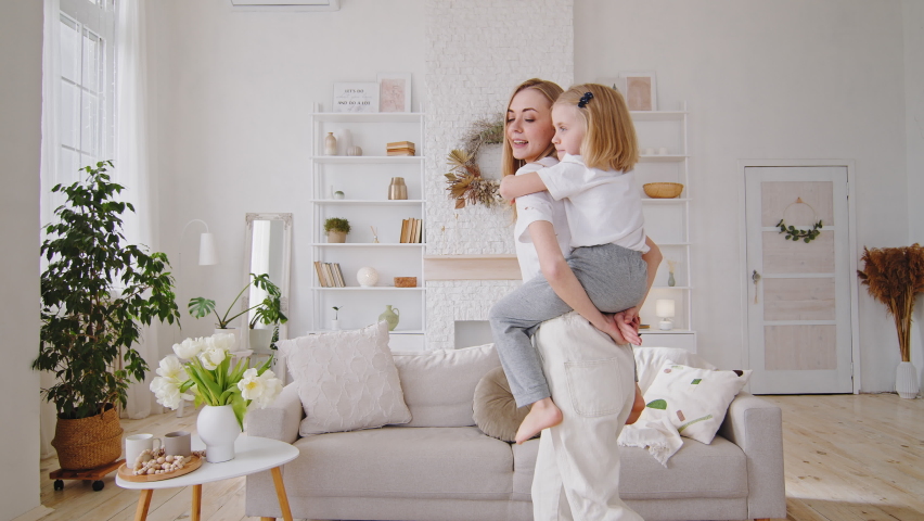 Caucasian mother babysitter adult woman blonde holding child girl on back carries baby piggyback playing with daughter small kid whirls rotates in circle at home in dancing living room having fun Royalty-Free Stock Footage #1073251418