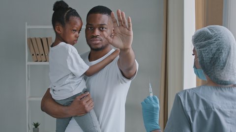 Young serious man afro american father holds in hands little African daughter child preschool girl puts palm in front of him refuses prohibits vaccination injection with vaccine to woman doctor nurse