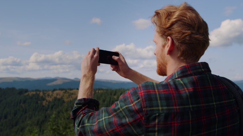 Serious man taking photos on mobile phone of picturesque landscape. Ginger guy making video on smartphone for blog. Back view male hiker spending weekend in mountains at summer  Royalty-Free Stock Footage #1073251997