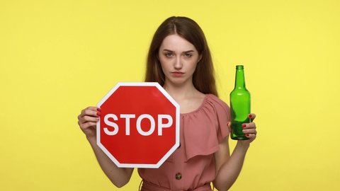 stop drinking alcohol.  Do not be drunk. Concentrated woman in elegant dress holding red stop sign and beer alcohol bottle, calls on to give up bad habit. Indoor, isolated on yellow background.
