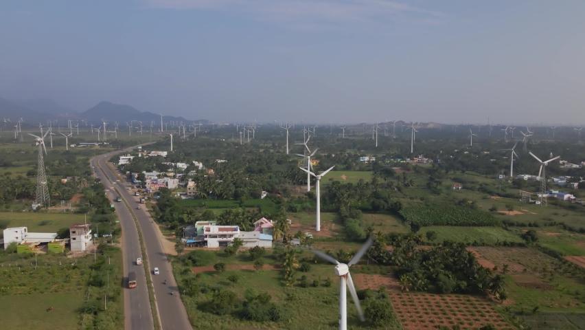A landscape wide drone aerial moving shot of a two-lane busy roadway dividing huge farm of wind turbines on a bright sunny day in India. sustainable, clean, and renewable energy concept  Royalty-Free Stock Footage #1073265842