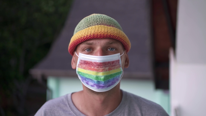 A young gay man looks at the camera close-up on his face mask painted under the LGBT flag.
