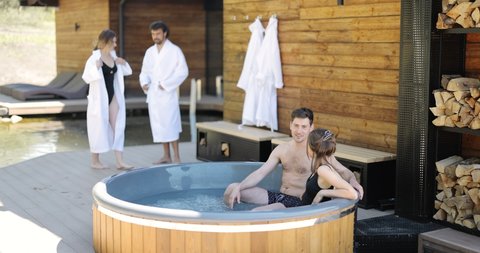 Company of friends bathing in outdoor jacuzzi or hot vat near the spa and lake. Couples have fun and relax in wellness center