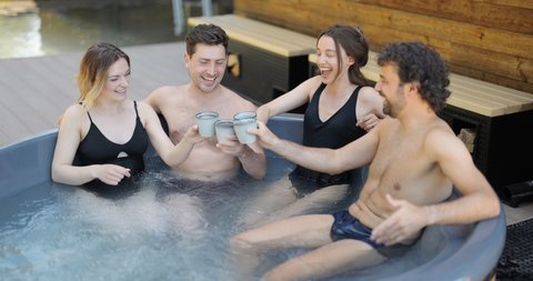 Company of friends bathing in outdoor jacuzzi or hot vat at the luxury spa. People relax and have fun drinking hot drinks in wellness center