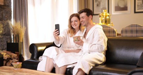 Woman and man in bathrobe sitting on sofa in living room of spa house and have a video call with friends on phone. Couple spending romantic time together in sauna