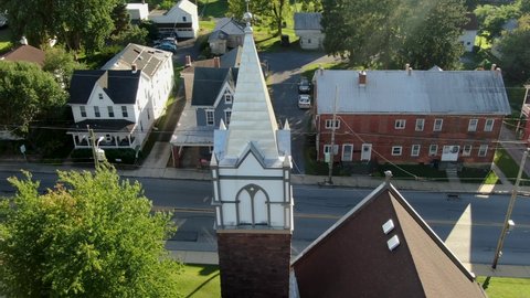 Aerial tilt reveals church steeple along road in old historic houses in USA, Small town America, August summer magic hour overhead view. Christianity Christian population theme.