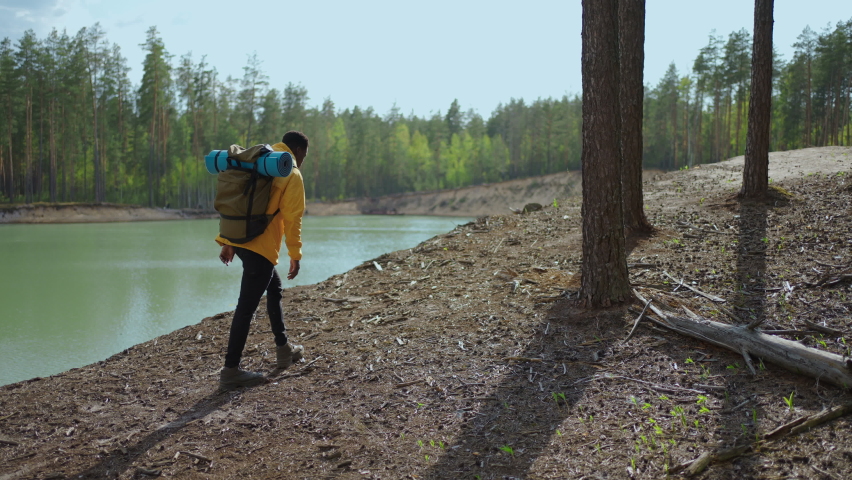 A young black man in a yellow cursor climbs up with a backpack uphill in the woods looking at the lake. Slow motion