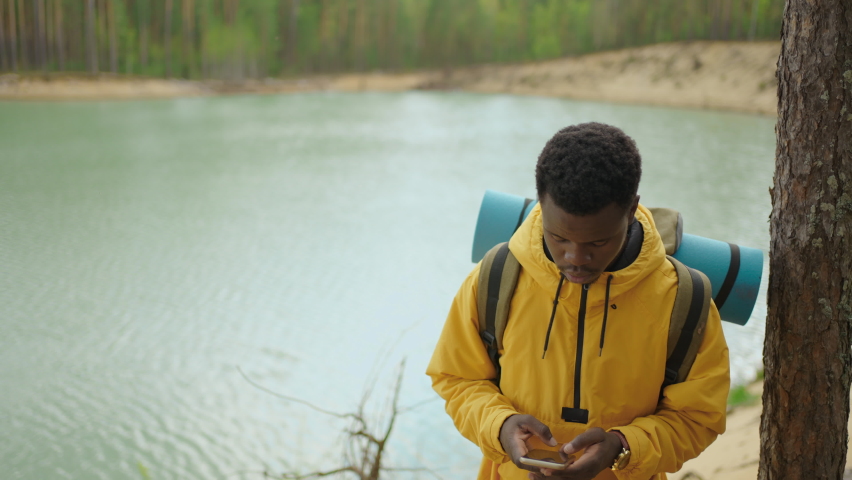 A black man is walking on top of a mountain and typing a message and photo on his smartphone. He has a large backpack on his shoulders. Hiking in the mountains. Top of the mountain