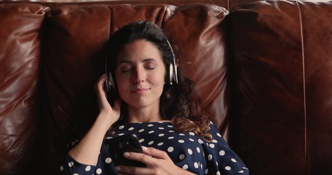 Head shot peaceful young beautiful woman resting on cozy sofa, listening slow music in wireless headphones, singing song with closed eyes, holding cellphone in hands, enjoying stress free weekend.