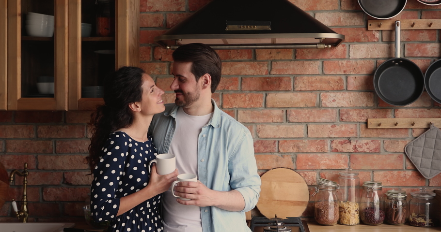 Loving young husband cuddling smiling beautiful wife in kitchen, enjoying pleasant conversation while drinking morning coffee together, spending leisure weekend lazy time communicating at own home. Royalty-Free Stock Footage #1073278145