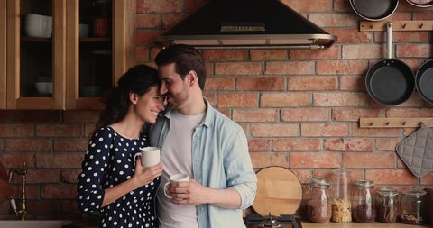 Loving young husband cuddling smiling beautiful wife in kitchen, enjoying pleasant conversation while drinking morning coffee together, spending leisure weekend lazy time communicating at own home.