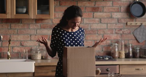 Confused young hispanic woman unwrapping cardboard box, feeling disappointed with received wrong or damaged item, displeased with negative online shopping experience or unreliable delivery service.