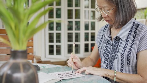 senior retired asian woman practicing writing Chinese calligraphy at home patio during free time