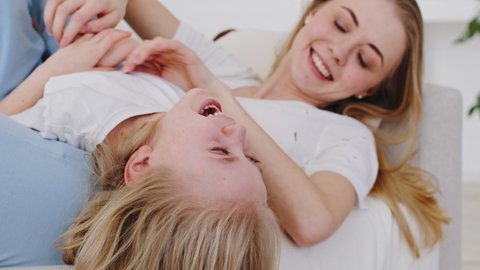 Close-up little girl child daughter weighs lies upside down with mother on sofa loving woman tickles baby playing game tickling laughs loudly having fun on couch, caucasian family spends time at home