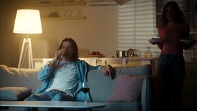 Restaurant service at home. Young relaxed man drinking wine, his positive woman bringing two portions of dinner to living room and sitting down on sofa, slow motion - Powered by Shutterstock - Get 15% off with code: PIKWIZARD15