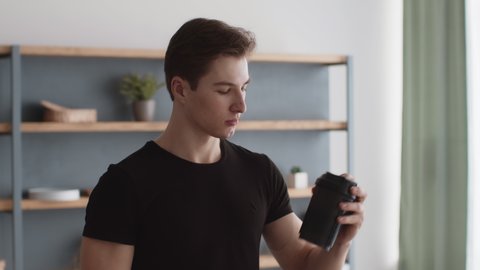 Weight gain diet. Young muscular man athlete mixing protein cocktail in sport shaker and drinking it, practicing sports workout at home interior, close up, slow motion