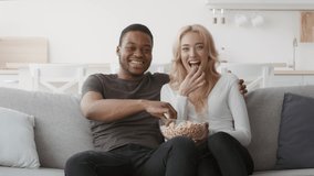 Multiracial Couple Laughing Watching TV Together Eating Popcorn And Having Fun Sitting On Couch At Home On Weekend, Looking At Camera. Weekend Leisure Concept. Zoom In