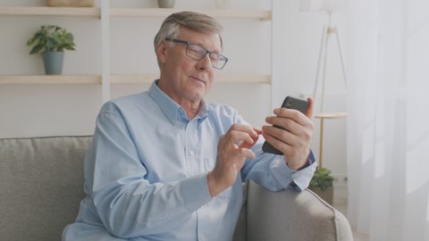 Smiling senior grey-haired man in eyeglasses sitting on couch and using mobile phone at home, chatting with his family or using mobile application, tracking shot, slow motion