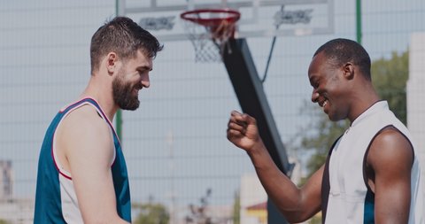 Sport and friendship. Multiethnic friends basketball players greeting each other at outdoor streetball court, hugging and bumping fists, side view, slow motion
