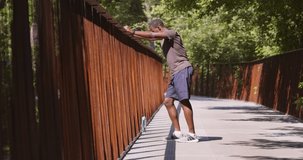 Break in sport workout. Sporty african american guy resting on bridge in urban park, standing at metal bars with water early in morning, slow motion
