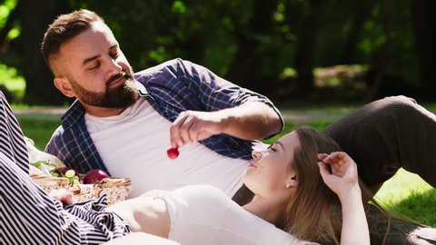 Romantic lunch in park. Close up portrait of happy affectionate couple enjoying picnic in green park, man feeding woman with fresh summer fruit, slow motion