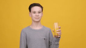 Cheerful Teen Boy Drinking Orange Juice And Smiling To Camera Posing With Glass Standing On Yellow Background. Studio Shot. Healthy Nutrition And Hydration For Teenager Guy Concept