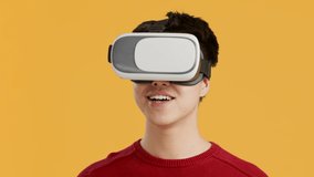 Happy Asian Millennial Guy Experiencing Virtual Reality Wearing VR Headset Posing Standing Over Yellow Studio Background, Looking At Camera. Augmented Reality Technology Concept