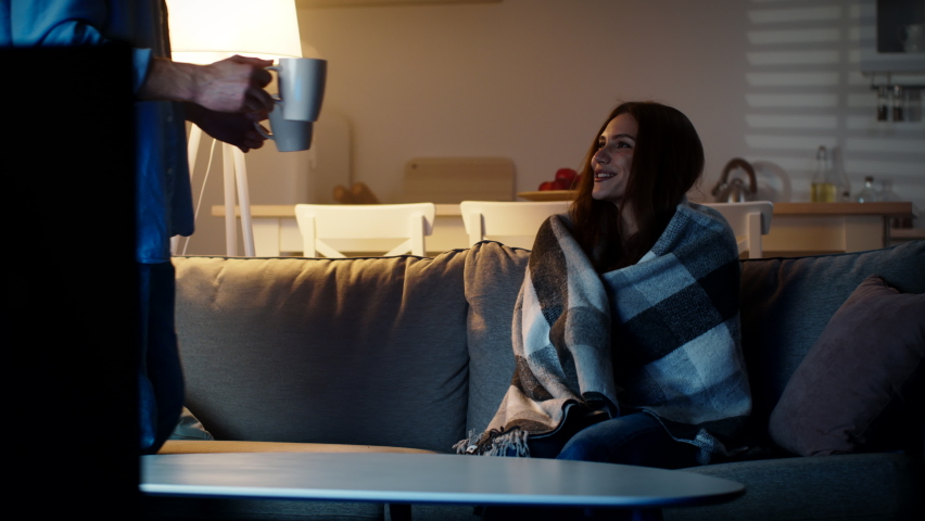 Love and care. Frozen woman sitting wrapped in plaid, her caring husband bringing two cups of tea and sitting down near her, watching tv in evening at home