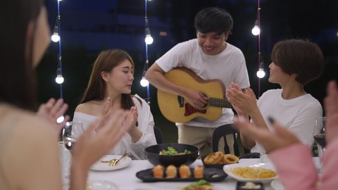 Group of Asian woman friends have outdoor dinner party with musician playing guitar in the garden at night. Beautiful female friendship enjoy celebration party eating food and drinking wine together.