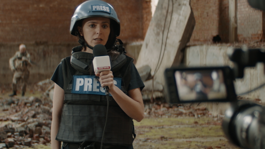 Behind the scenes of female war journalist correspondent wearing bulletproof vest and helmet reporting live near destroyed building, military personnel in the background