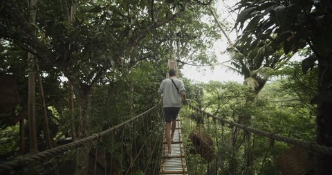 Dolly view following a man carefully walking across a shaking hanging bridge in a tropical rainforest in Bali, Indonesia