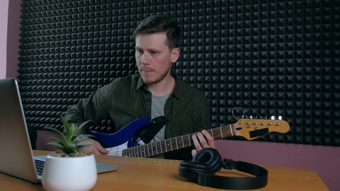 European Man Learning an Electro Guitar in Music Studio with laptop. E-learning, Online Education Concept. Male self education online. Guy plays guitar front of laptop. 