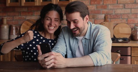 Overjoyed young loving family couple looking at smartphone screen, getting message with good news, celebrating online lottery gambling giveaway win, receiving low price discount shopping promo code.