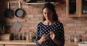 Relaxed dreamy young latin caucasian woman standing in kitchen, using smartphone application, web surfing interesting dishes receipt or communicating distantly in social network with friends at home.