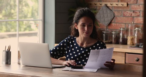 Pleasant young hispanic woman sitting at table with computer, calculating paper bills, managing bank loan mortgage payments, planning monthly budget, paying taxes, satisfied with enough money.