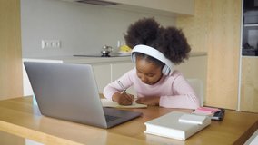 Little african american girl wearing headset studying from home answering during online school lesson writing in copy-book. Mixed-race schoolgirl enjoy homeschooling. Distance education, e-learning