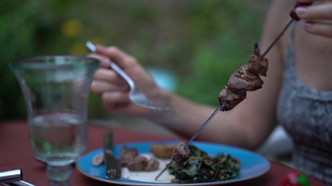 Closeup of woman holding a skewer as she pulls meat off a skewer onto her plate outside.