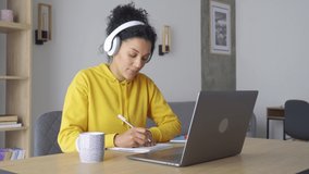 African American mixed race adult student wearing headphones having virtual meeting online class with teacher, writing notes. Webinar chat at home office. Video e learning conference call on pc.