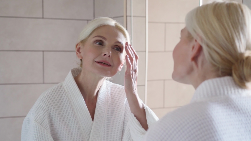 Attractive beautiful mid age adult 50 years old blonde woman standing in bathroom touching face with hands, smiling to reflection in mirror doing daily morning beauty routine. Older skin care concept. Royalty-Free Stock Footage #1073342342