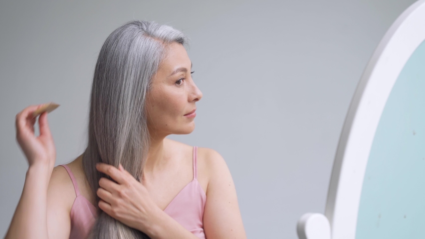 Senior attractive middle 50 years aged asian woman with gray hair looking at mirror reflection combing tangled gray hair. Alopecia hair loss prevention treatment after menopause advertising concept. Royalty-Free Stock Footage #1073342360