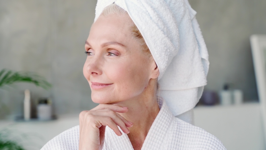 Happy smiling attractive middle aged woman wearing bathrobe and white towel touching face looking at camera. Advertising of skin care spa procedures concept. Closeup portrait. | Shutterstock HD Video #1073342369
