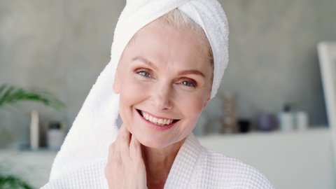 Happy smiling attractive middle aged woman wearing bathrobe and white towel touching face looking at camera. Advertising of skin care spa procedures concept. Closeup portrait.