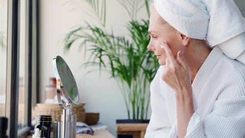 Attractive mid age older adult 50 years old blonde woman wears bathrobe and towel in bathroom applying nourishing antiage face skin care cream treatment, looking at mirror doing daily beauty routine.