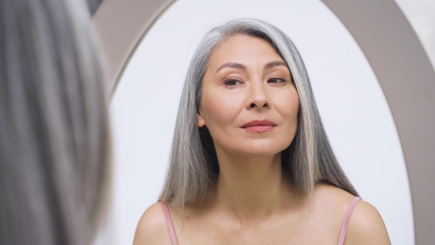 Senior older middle aged Asian woman with grey hair with radiant face with perfect skin looking at mirror smiling to her reflexion touching massaging skin. Ads of senior dry skin care treatment. Royalty-Free Stock Footage #1073342411