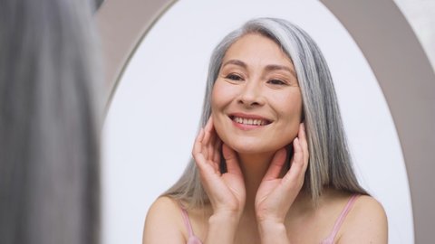 Senior older middle aged Asian woman with grey hair with radiant face with perfect skin looking at mirror smiling to her reflexion touching massaging skin. Ads of senior dry skin care treatment.