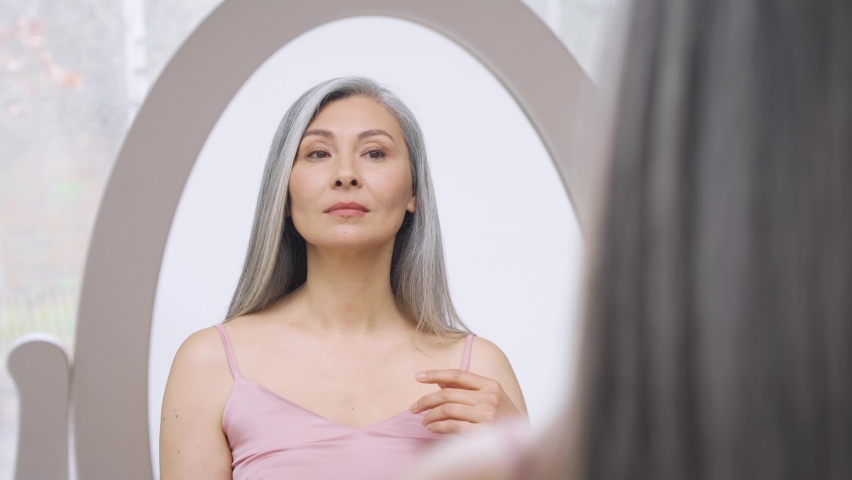 Senior older middle aged Asian woman with grey hair with radiant face with perfect skin looking at mirror smiling to her reflexion. Ads of makeup foundation for natural glow skin. Royalty-Free Stock Footage #1073342417
