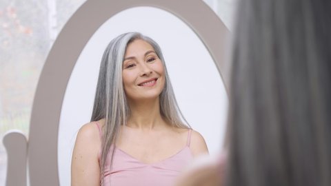 Senior older middle aged Asian woman with grey hair with radiant face with perfect skin looking at mirror smiling to her reflexion. Ads of makeup foundation for natural glow skin.