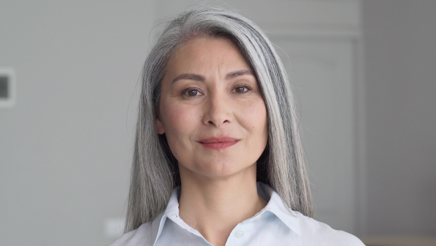 Happy smiling stylish confident 50 years old Asian female professional standing looking at camera at gray background. Portrait of sophisticated grey hair woman advertising products and services. | Shutterstock HD Video #1073342420
